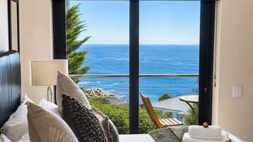 A bedroom with a large window overlooking the sea, a bed with multiple pillows, a bedside lamp, and a chair with towels.