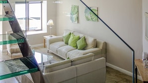 A modern living room with a glass staircase, a sofa set, and a bed with a tufted headboard.