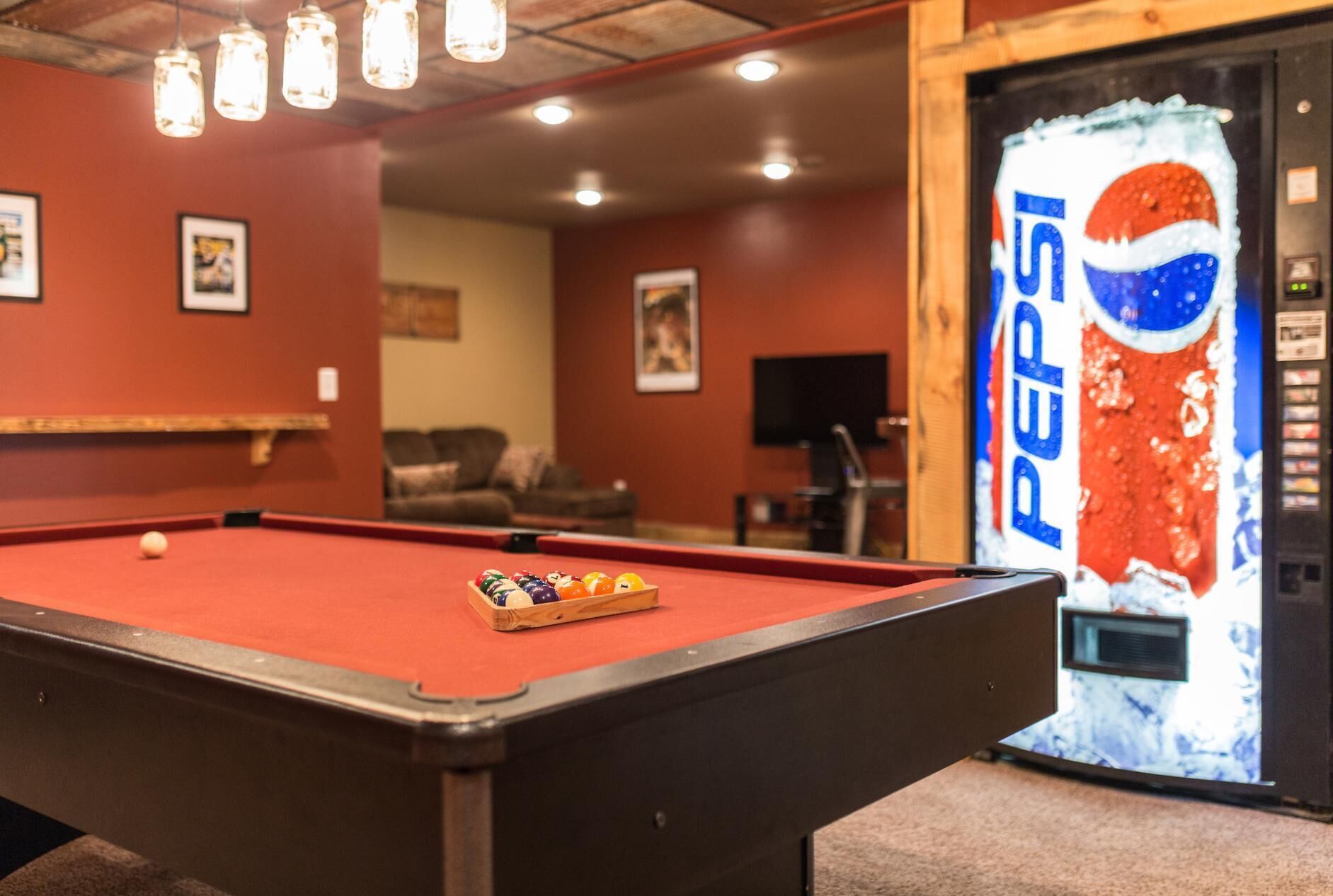 A pool table in a room with a large Pepsi refrigerator, a couch, and a television in the background.