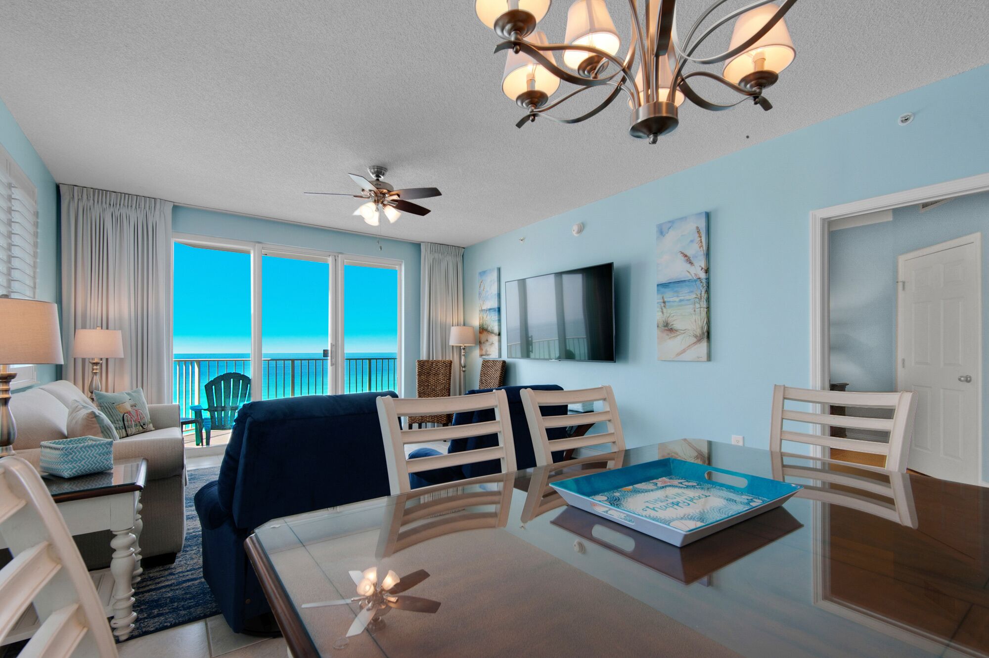 A hotel room with a balcony, a ceiling fan, a dining table with chairs, a TV, and a painting on the wall.