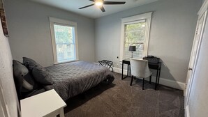 Un dormitorio con cama, escritorio y silla. Hay dos ventanas con persianas, un ventilador de techo y piso alfombrado.