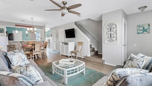 Ein geräumiges Wohnzimmer mit Deckenventilator, einer Essecke mit Holztisch und -stühlen, einer Treppe und einem an der Wand befestigten Fernseher.