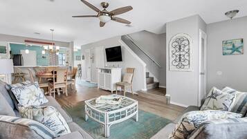 A spacious living room with a high ceiling fan, a dining area with wooden furniture, a staircase, and a TV mounted on the wall.