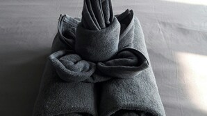 Una cama bien hecha con edredón gris, una toalla enrollada y un cojín sobre una cama con cabecera capitonada.