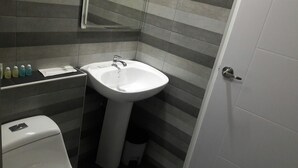 Un baño con lavamanos blanco, un espejo encima y un inodoro. Hay artículos de aseo en la encimera.