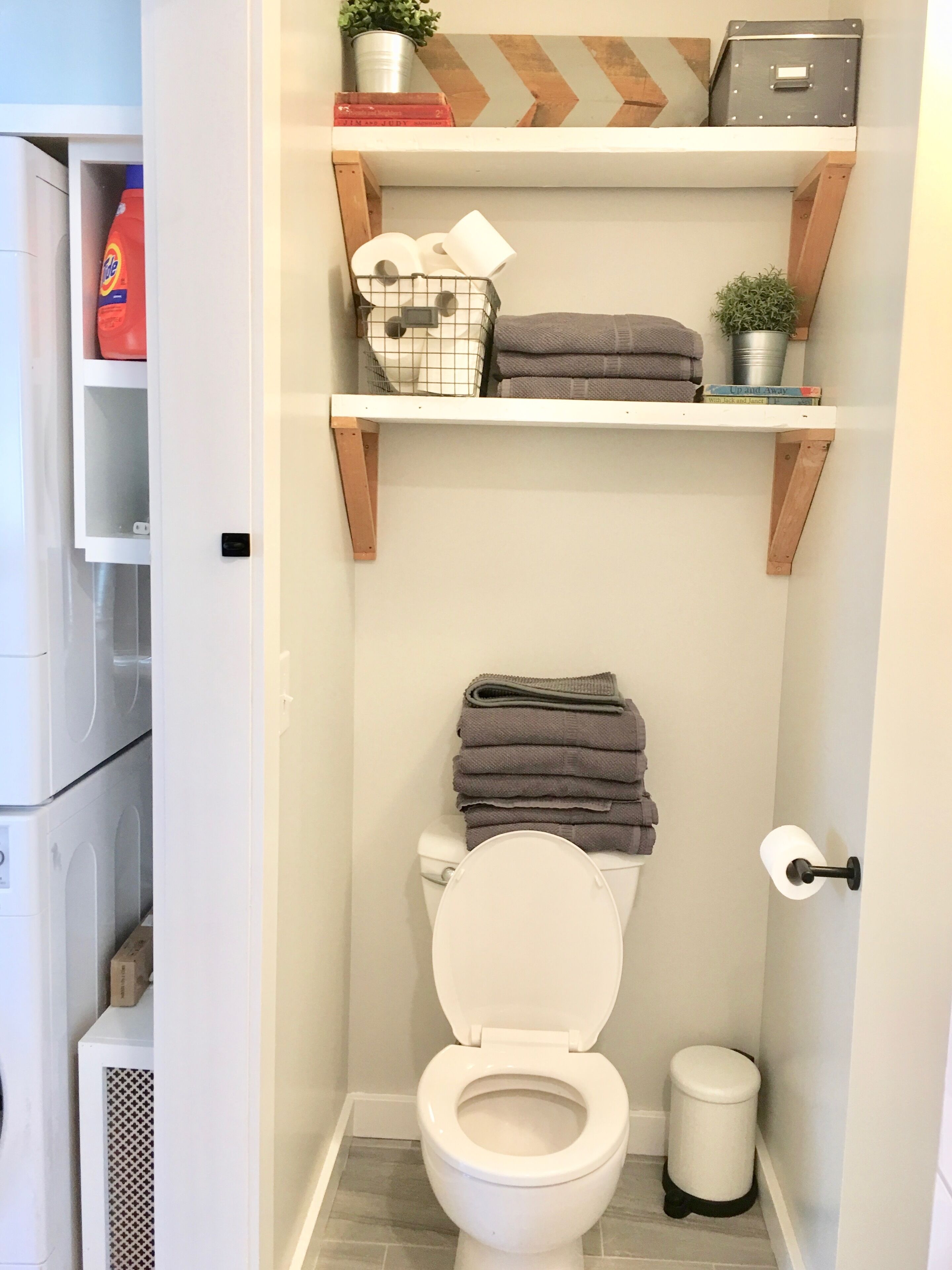 A compact bathroom with a toilet, a trash bin, and shelves above the toilet holding towels and a laundry basket.