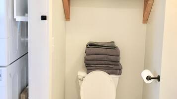 A compact bathroom with a toilet, a trash bin, and shelves above the toilet holding towels and a laundry basket.