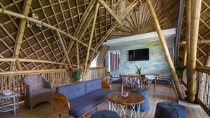 A spacious lounge with a bamboo roof, comfortable seating, a stone wall with a TV, and a wooden floor. - Wadari Retreat Villa Ubud (Ubud)