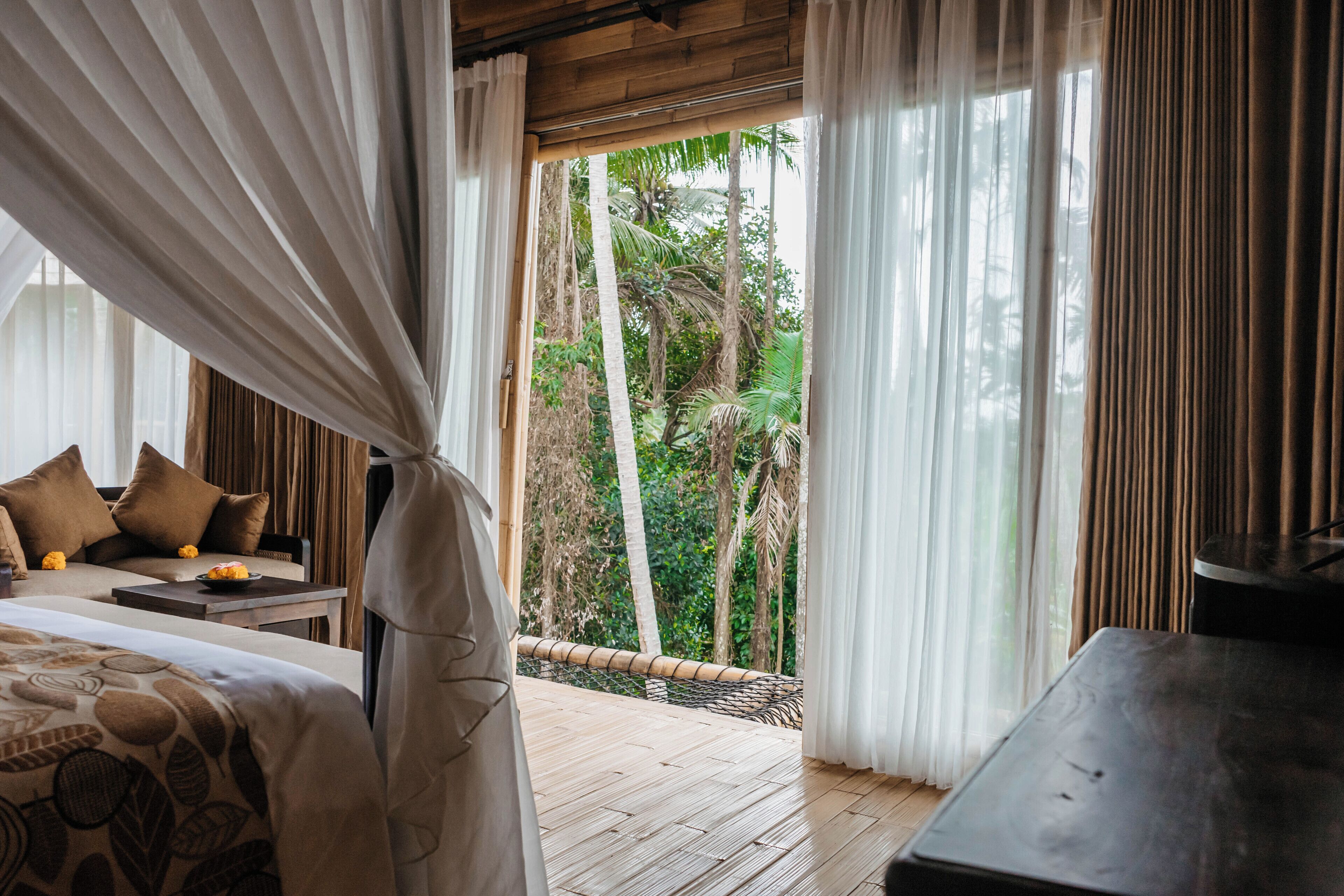 Ein Schlafzimmer mit einem Bett, einem Holztisch und Blick ins Freie durch transparente Vorhänge.