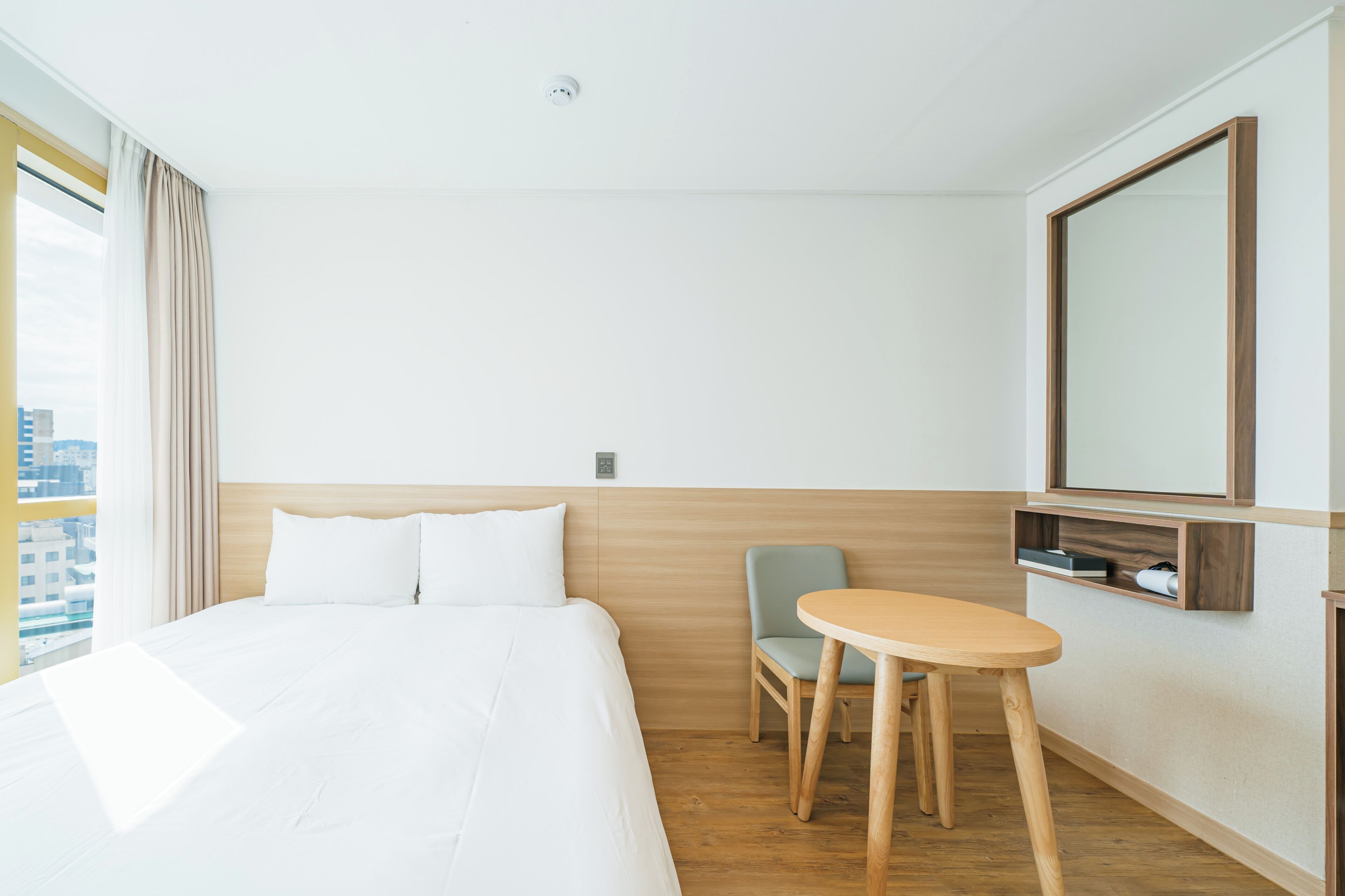 Habitación de hotel con cama, una silla, una mesita, un espejo y ventana con cortinas.