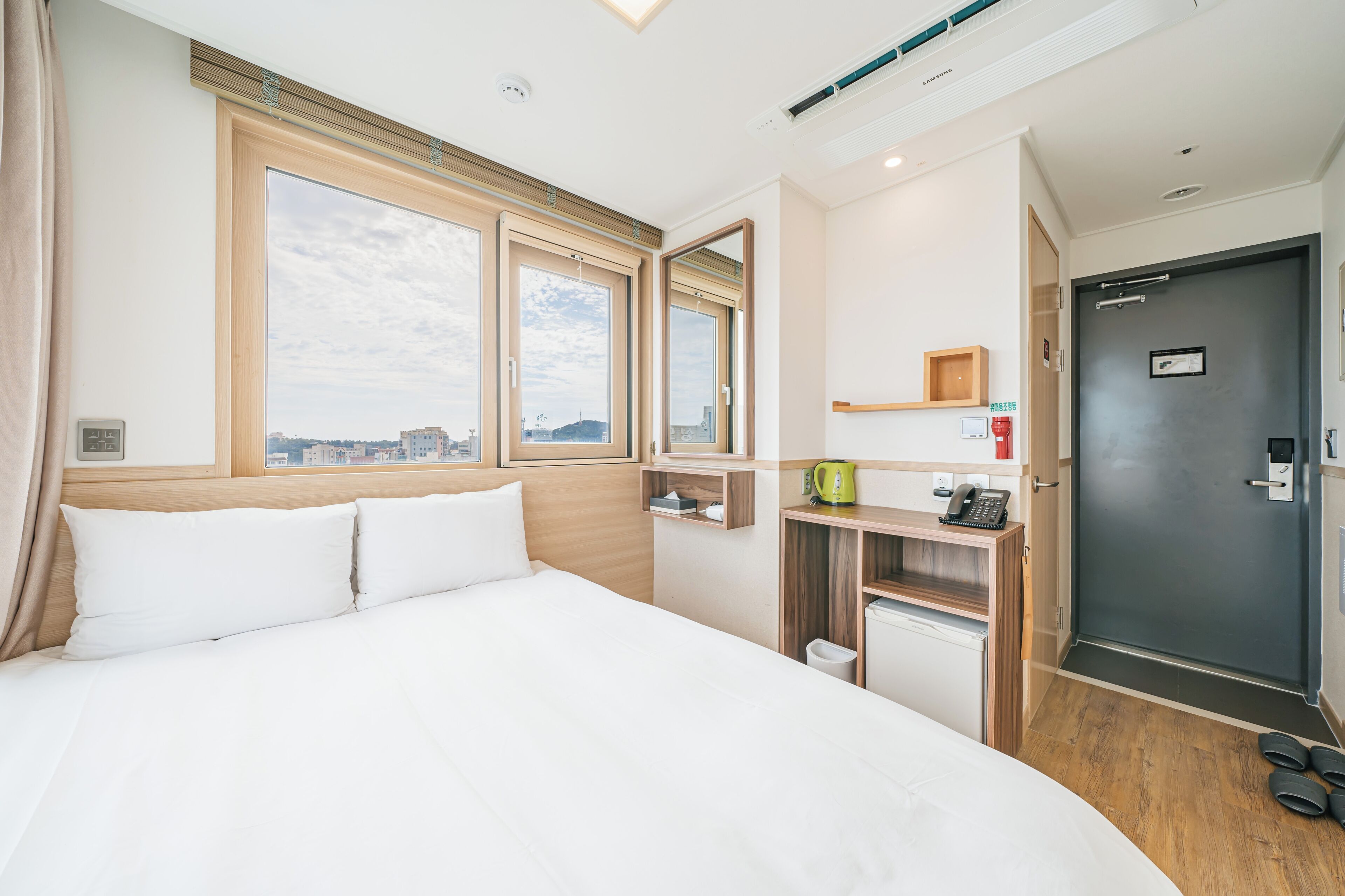 Habitación de hotel con una cama grande, un escritorio con teléfono, un refrigerador pequeño y vista a la ciudad a través de amplias ventanas.
