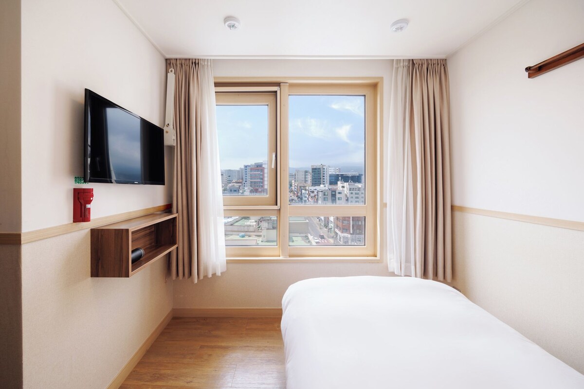 Habitación de hotel con una cama, un televisor montado en la pared, una ventana con cortinas y vistas a la ciudad.