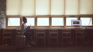 Eine Person sitzt an einem Tisch mit einem Koffer, einem Computerbildschirm und einem Fenster mit Jalousien.