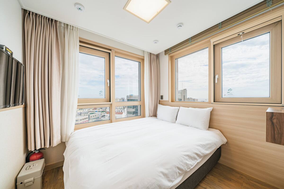 Een hotelkamer met een groot bed, twee ramen, een plafondlamp en uitzicht op de stad.
