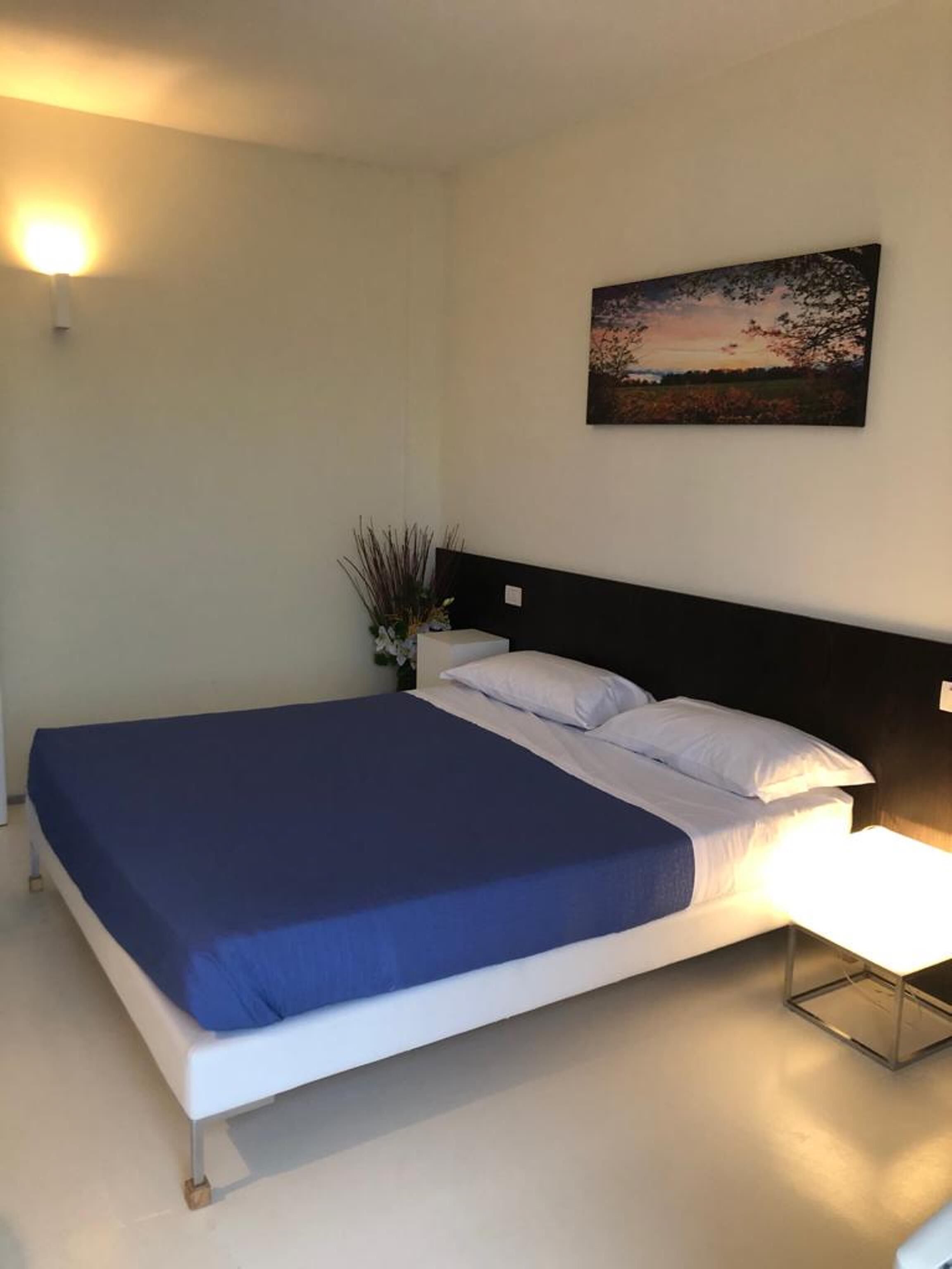 A modern bedroom with a large bed, a nightstand, a wall-mounted lamp, and a painting of a landscape.