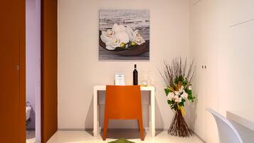 A modern, well-lit room with a dining area featuring a white table, an orange chair, and a green rug. A painting of white flowers is on the wall, and a vase with flowers is on the table.