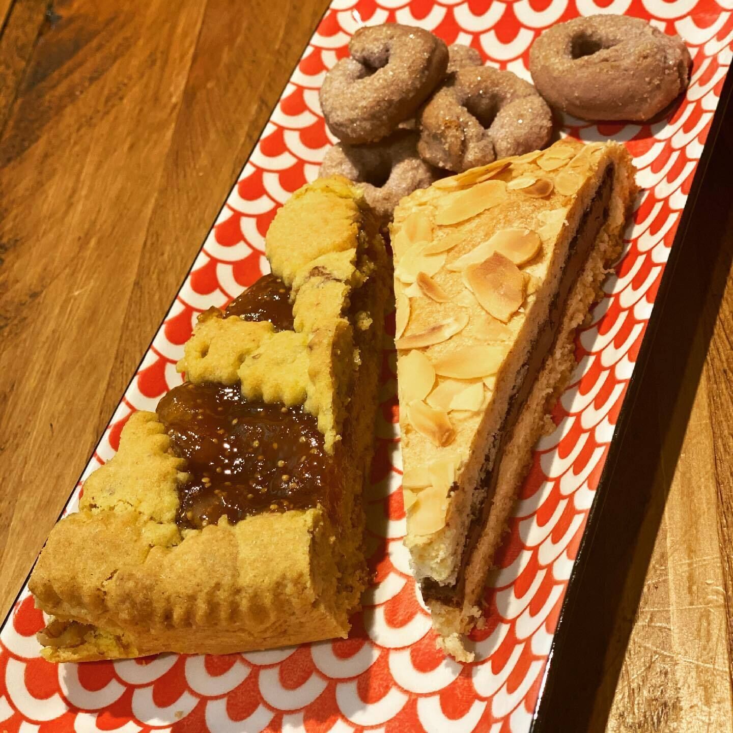 A rectangular plate with a red and white pattern, containing a slice of pie with a golden crust and a dark filling, a slice of pie with a flaky crust and a light filling, and three round, sugar-coated pastries.