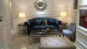 Een woonkamer met een blauwe bank, een witte poef, twee bijzettafels met lampen, en een decoratieve spiegel aan de muur.