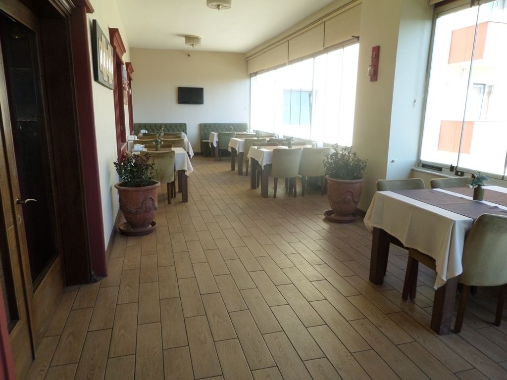 A restaurant with wooden flooring, tables with white tablecloths, potted plants, and large windows allowing natural light.