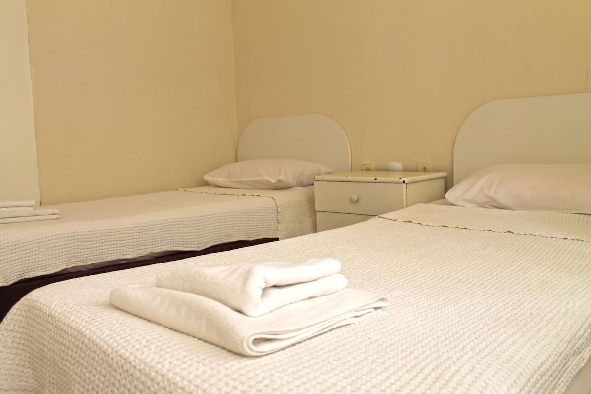 A hotel room with two beds, each with a white headboard and a white blanket. There is a nightstand with a lamp and a small table between the beds.