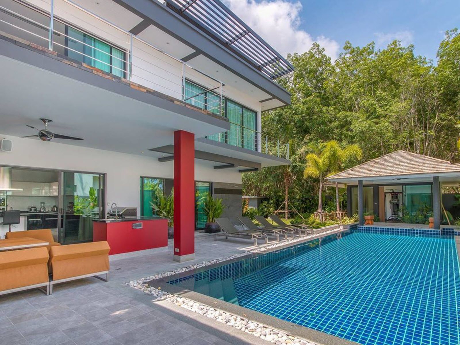 A modern house with a swimming pool, a red pillar, and a covered outdoor area with lounge chairs.