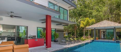 A modern house with a swimming pool, a red pillar, and a covered outdoor area with lounge chairs.