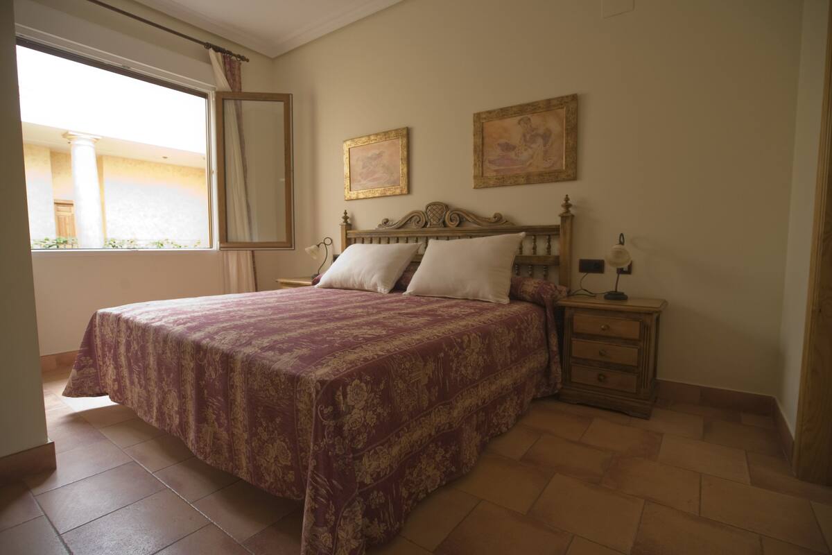 Un dormitorio con una cama grande, una mesita de noche, dos cuadros enmarcados en la pared y una ventana con cortinas.