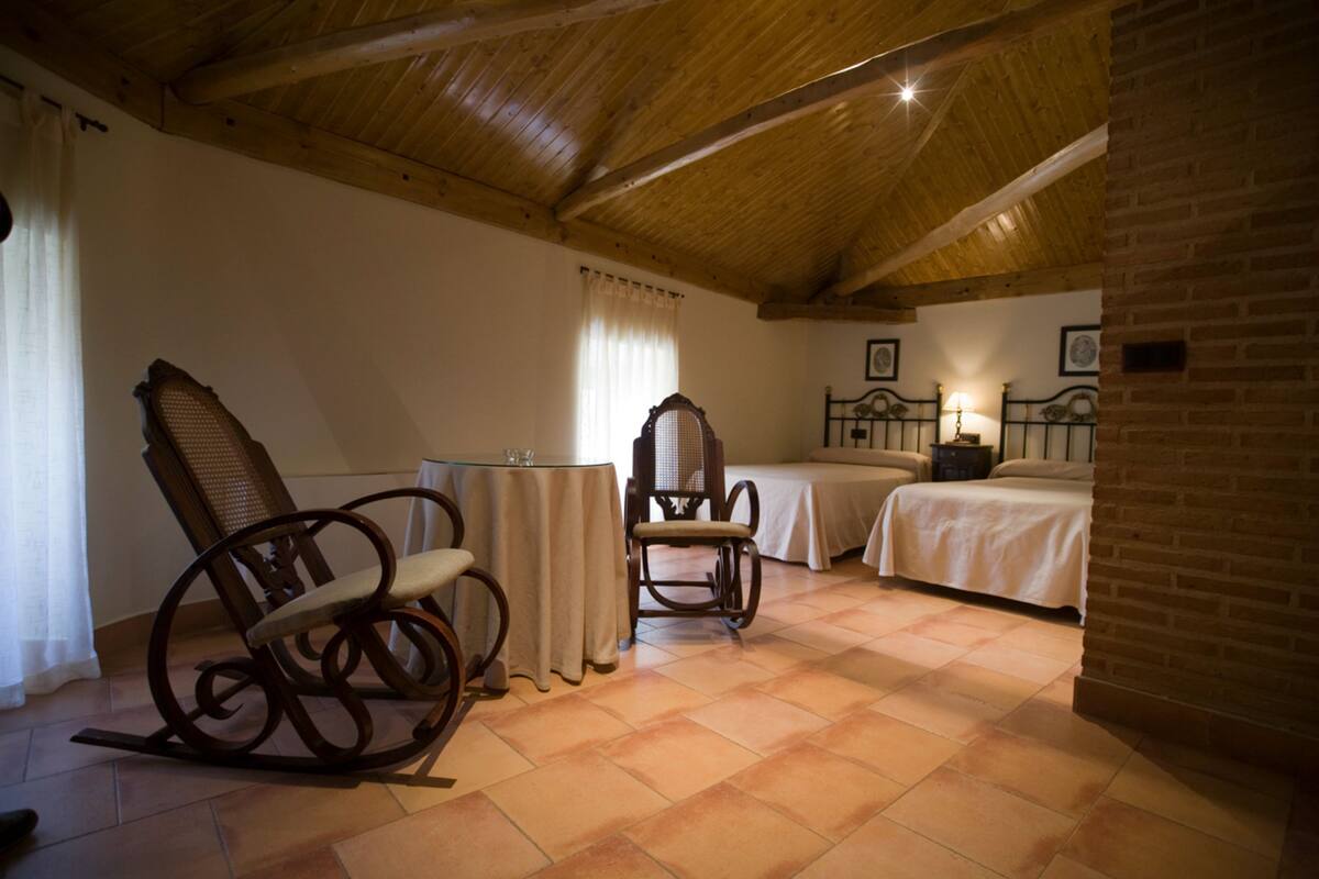 Un dormitorio con techo de madera, una mecedora, una mesita de noche y una cama con cabecera.