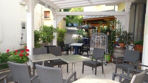 Ein Außenbereich mit Terrasse, Korbmöbeln, Topfpflanzen und Blick auf die Straße im Hintergrund.