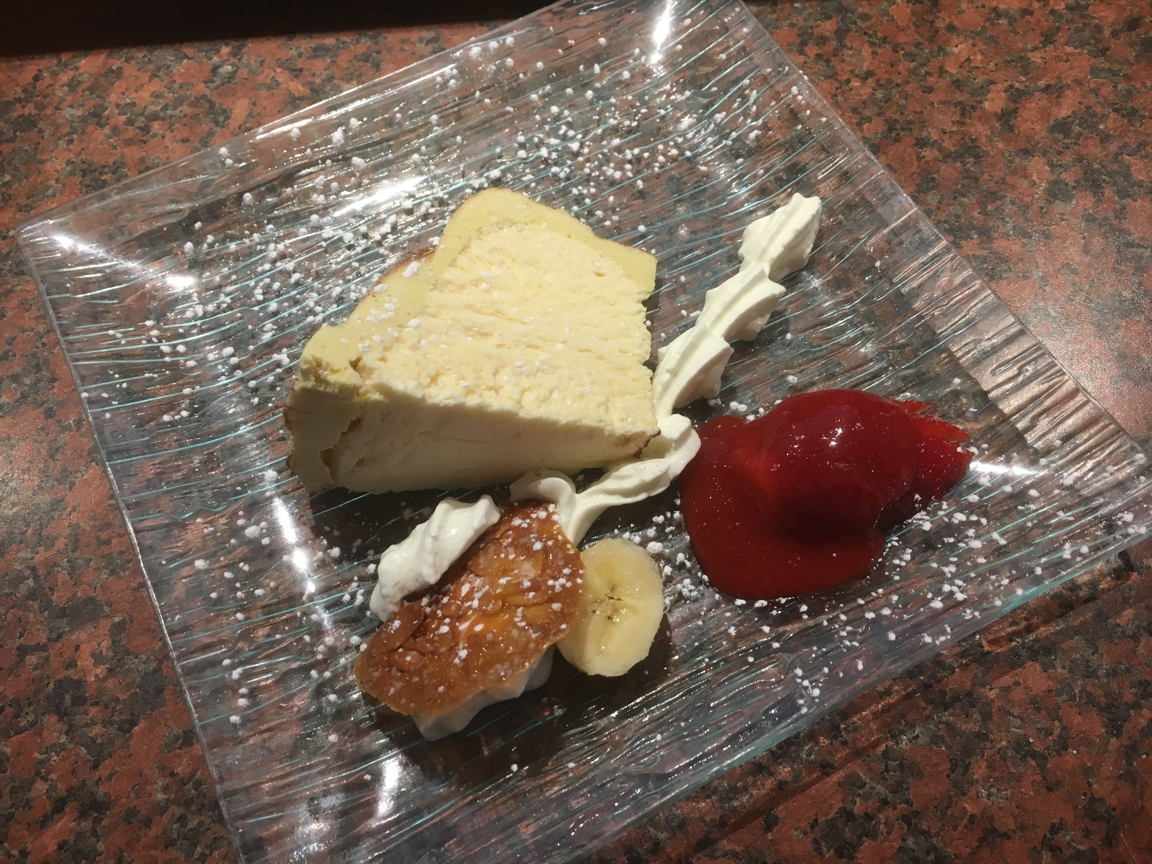 Un postre sobre un plato de vidrio transparente con crema batida, una porción de pastel, una cereza y un trozo de fruta.