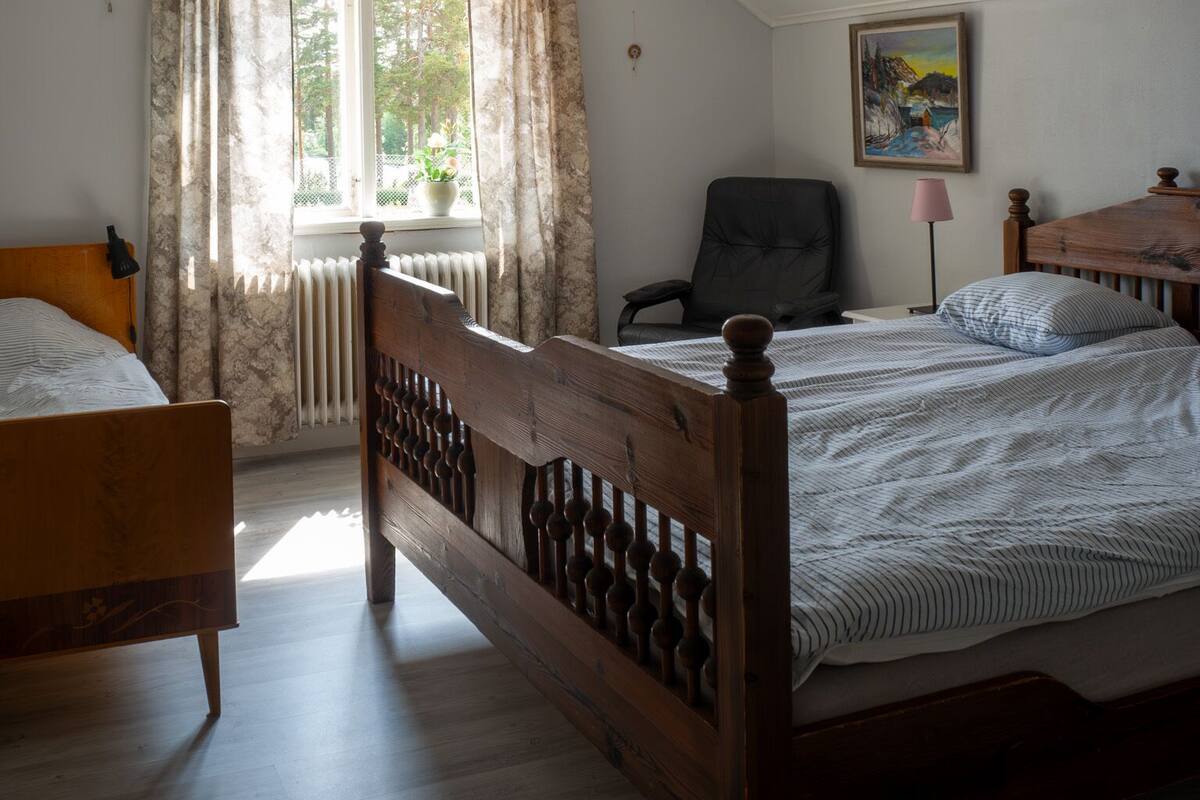 Quarto com cama de madeira, uma cadeira, uma escrivaninha, um radiador e janela com cortinas.