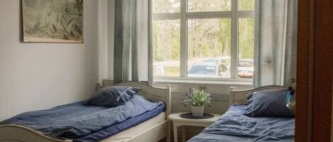 A room with two single beds, a window with curtains, a small table with a plant, and a painting on the wall.