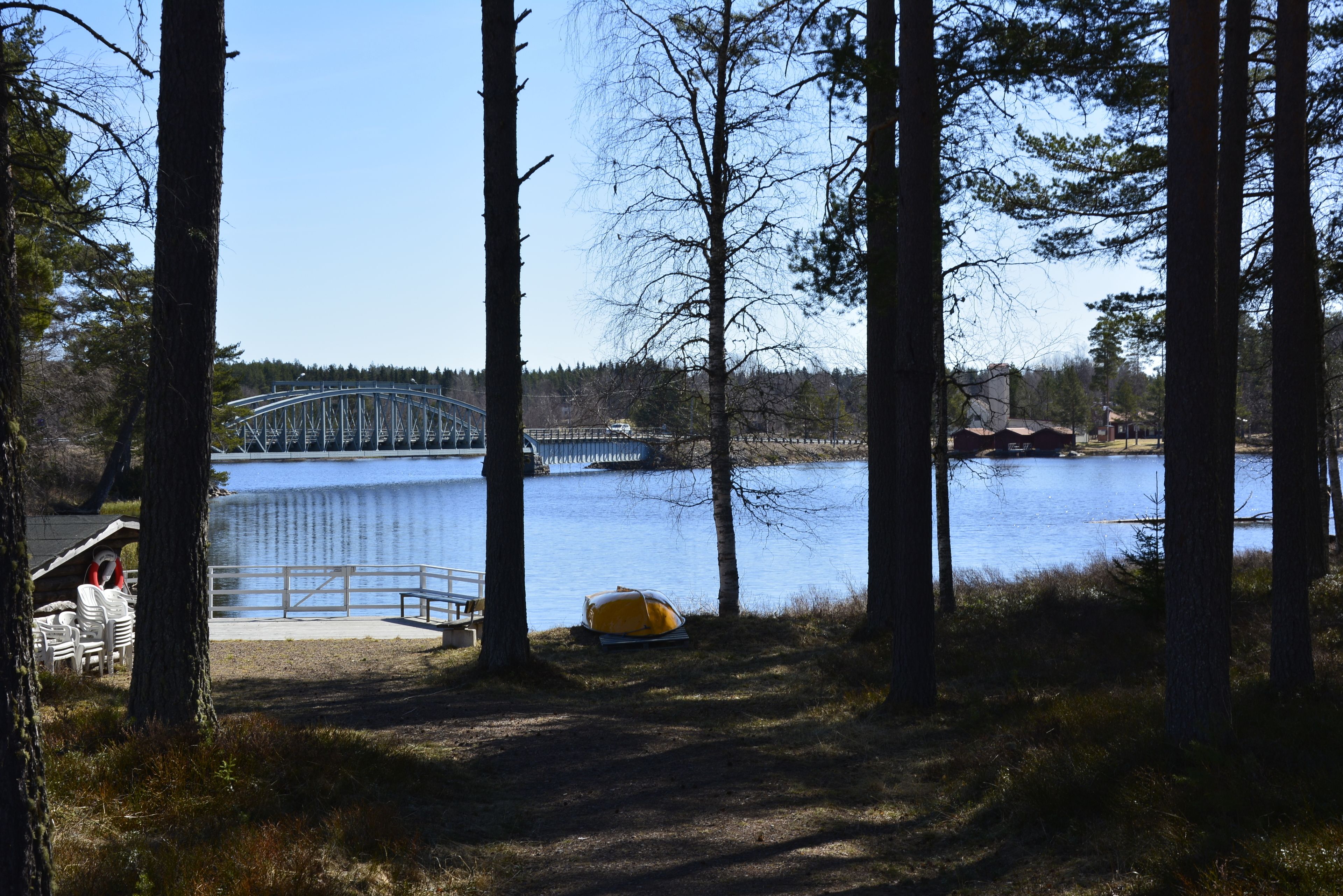 A lakeside view with a bridge, trees, and a camping area with chairs and a tent.