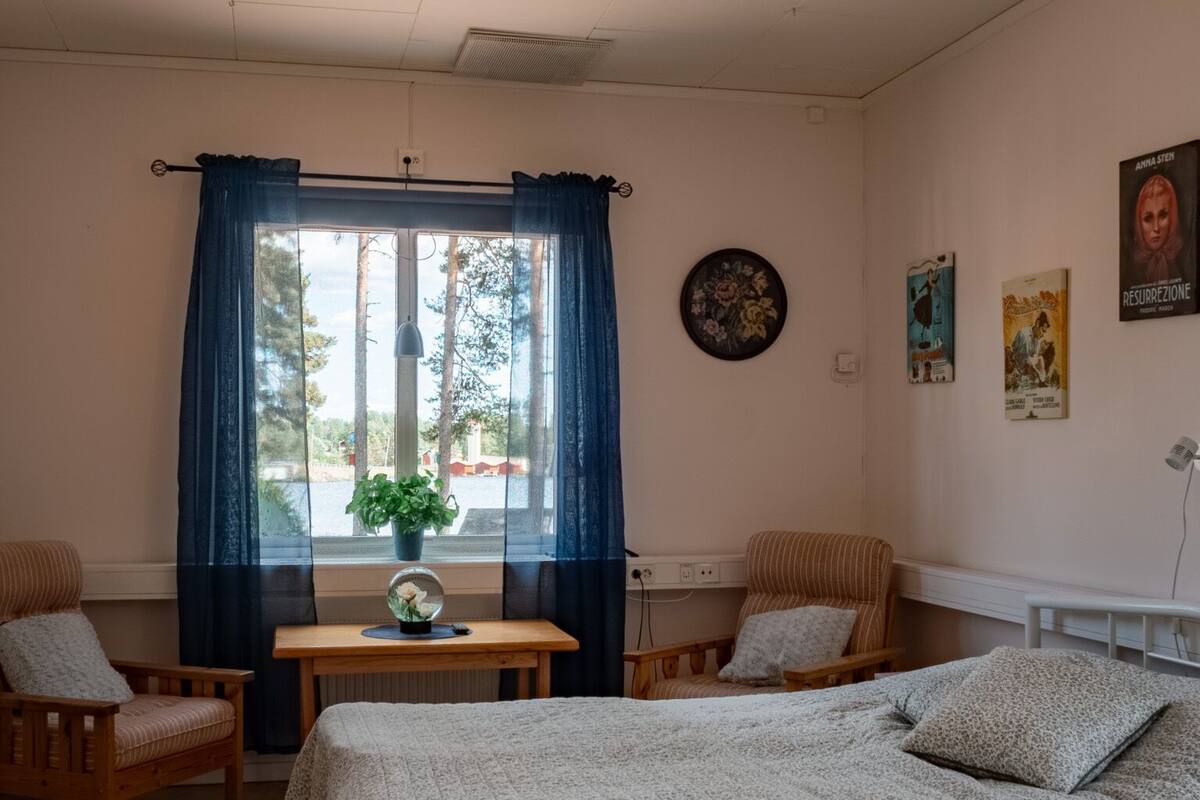 Quarto com cama, duas cadeiras, uma mesinha e janela com cortinas.