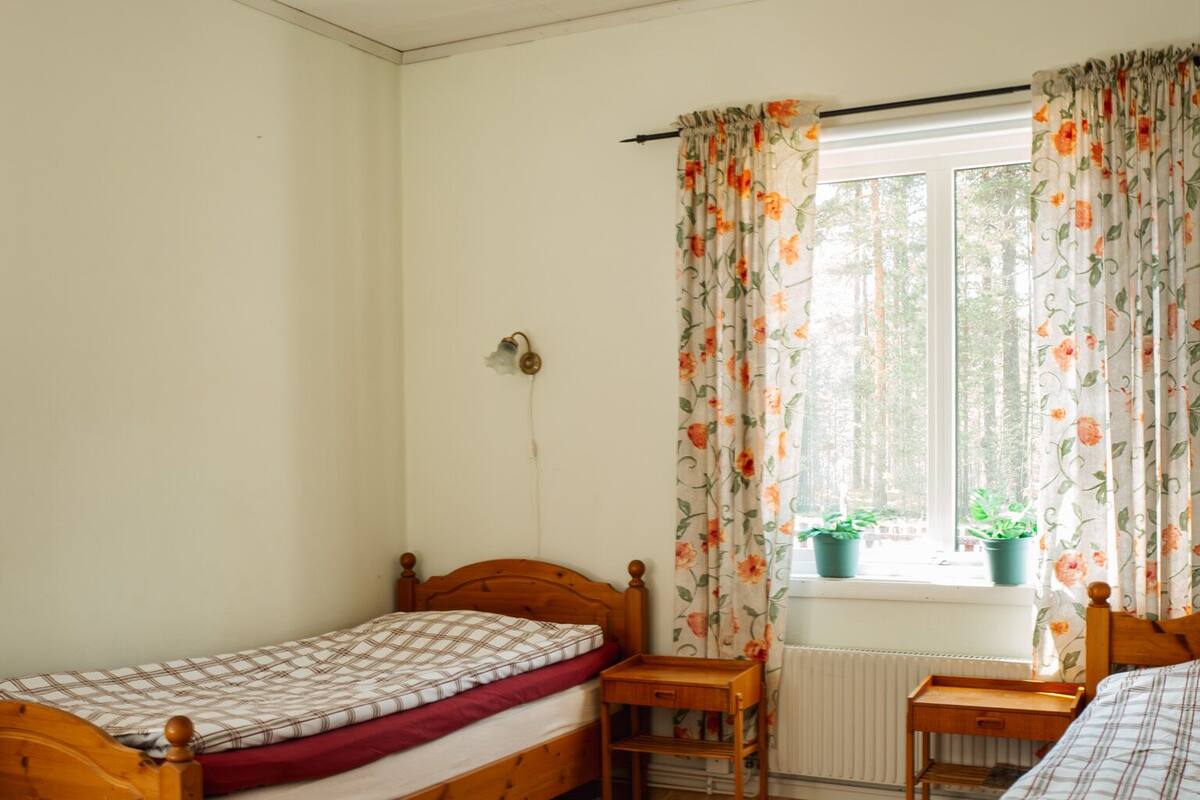 Quarto pequeno e bem iluminado com uma cama de madeira, uma pequena escrivaninha e uma janela com cortinas florais.