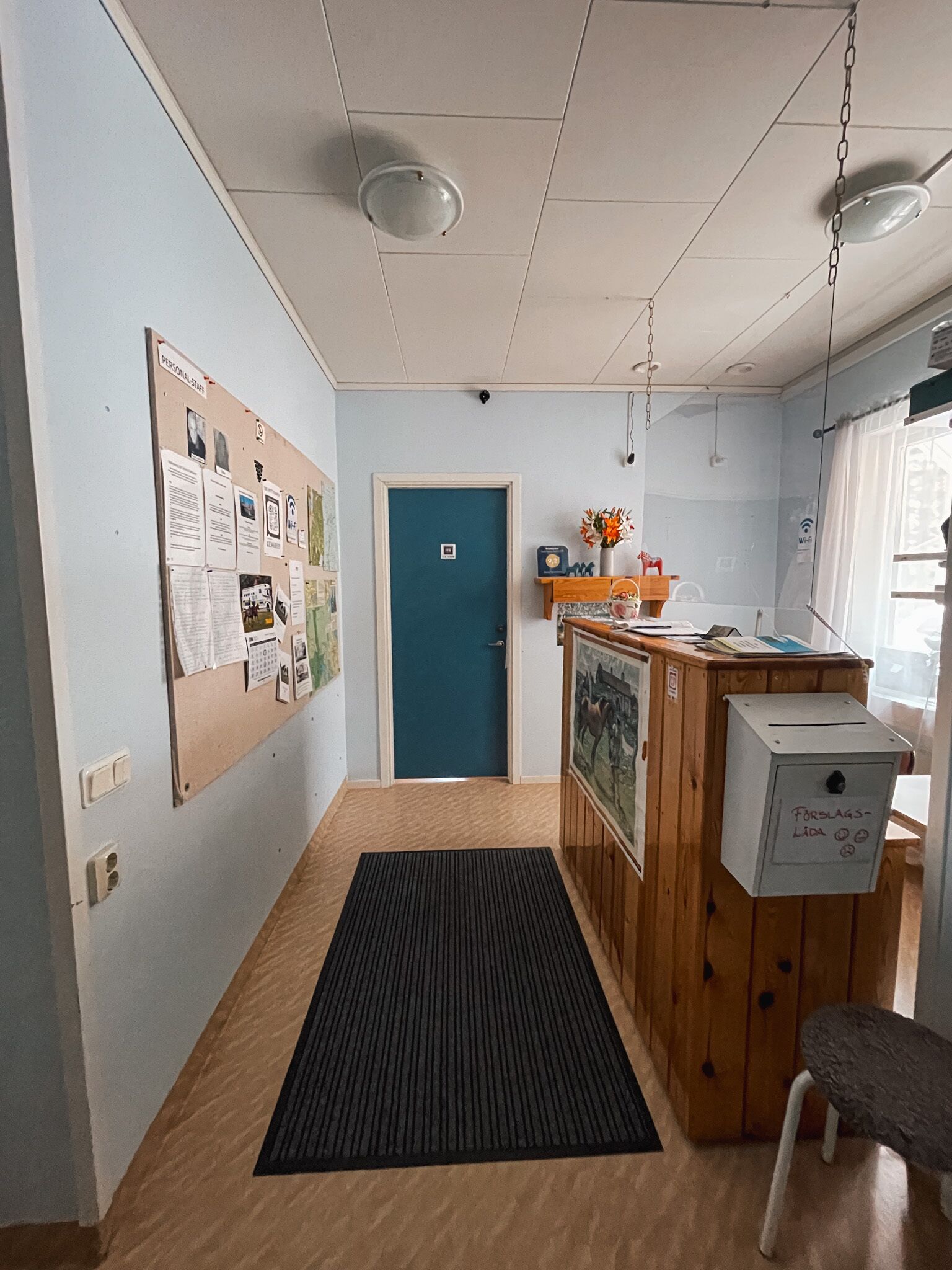 A hallway with a bulletin board, a blue door, a wooden counter with a cash register, and a small stool.