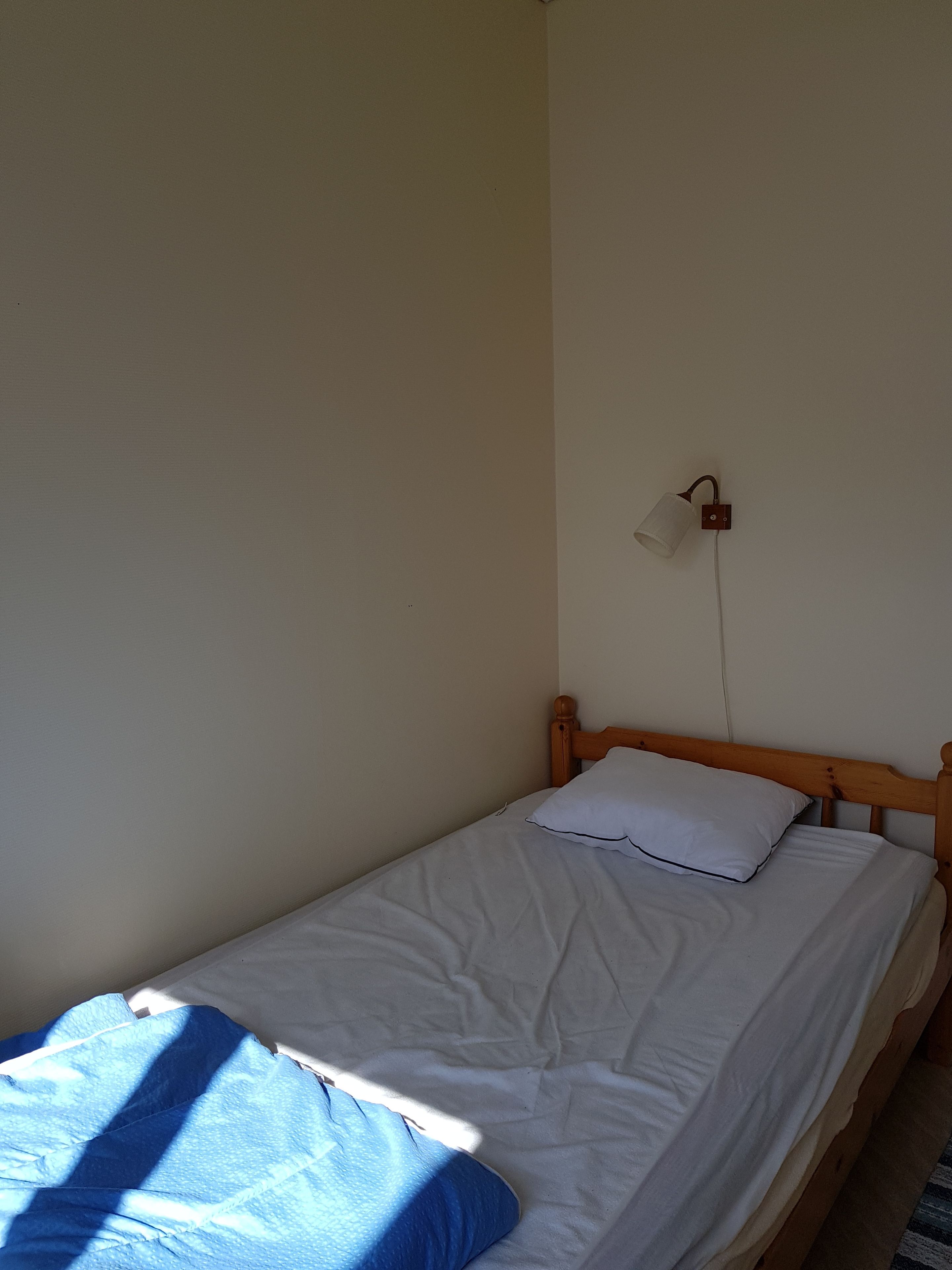 A single bed with a wooden headboard, white bedding, and a white lamp on the wall.
