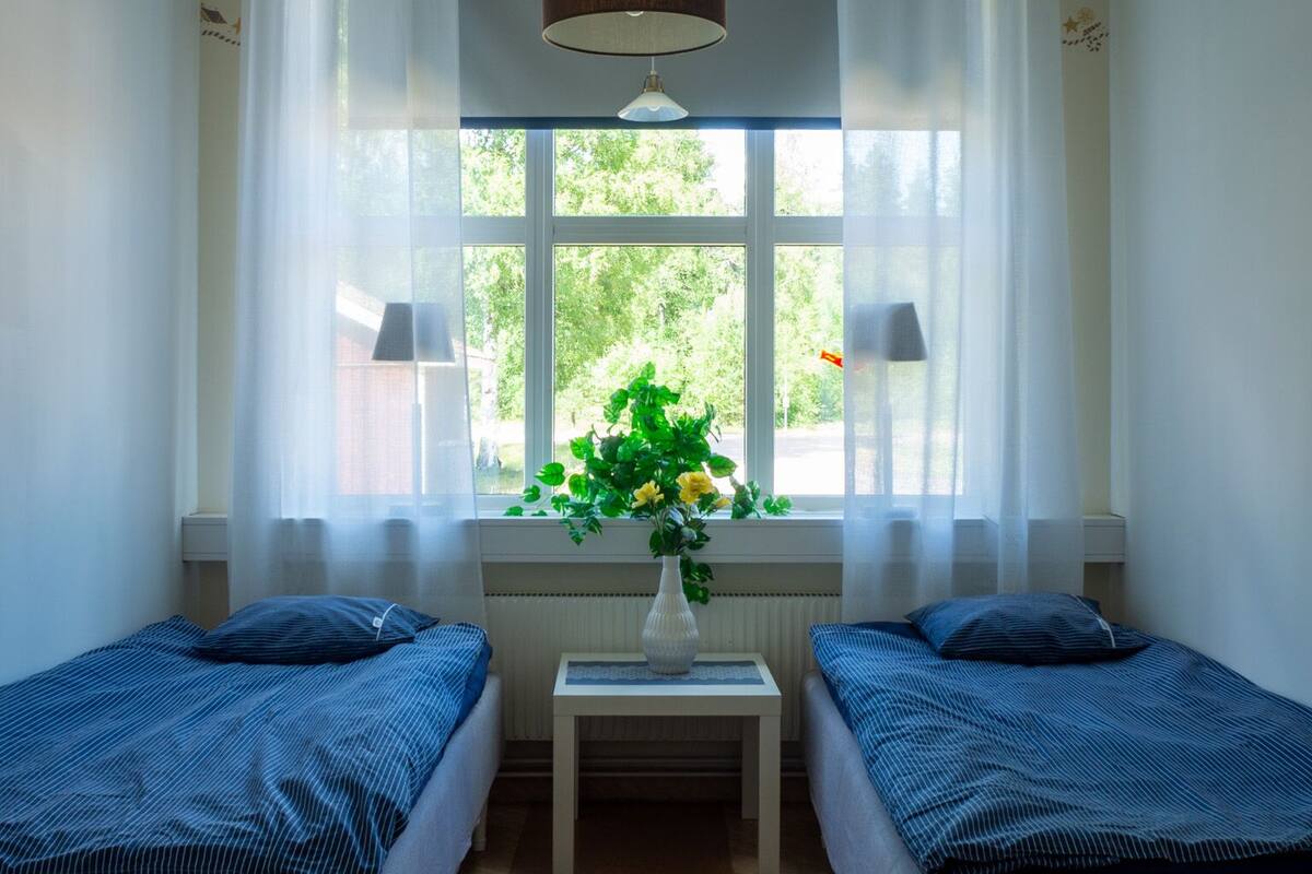 Quarto com duas camas de solteiro, uma mesinha com um vaso de flores e uma janela com cortinas.