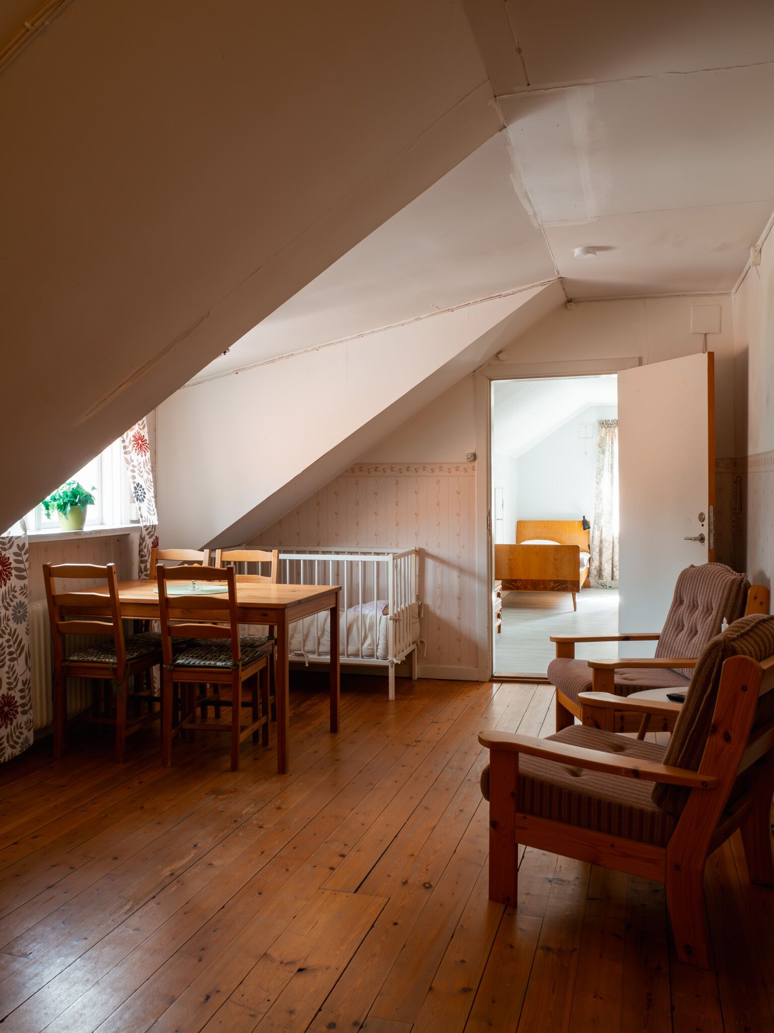 A cozy attic room with a wooden floor, a dining table with chairs, a crib, and a bed.