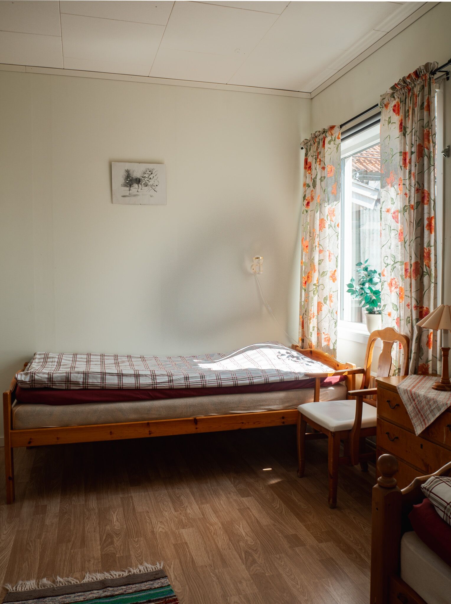 Quarto pequeno com uma cama de solteiro, estrutura de madeira, uma cadeira, uma mesinha e uma janela com cortinas florais.