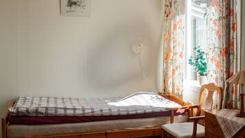Quarto pequeno com uma cama de solteiro, estrutura de madeira, uma cadeira, uma mesinha e uma janela com cortinas florais.