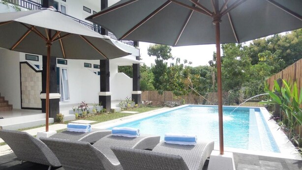 Piscina con sillas de descanso y sombrillas, rodeada por un jardín y un edificio.