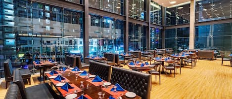 Un restaurante moderno con varias mesas, asientos tipo banqueta de cuero negro y grandes ventanales que ofrecen vistas a la ciudad.
