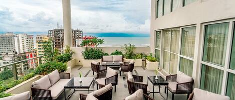 A balcony with wicker furniture, a view of the sea, and city buildings in the background.