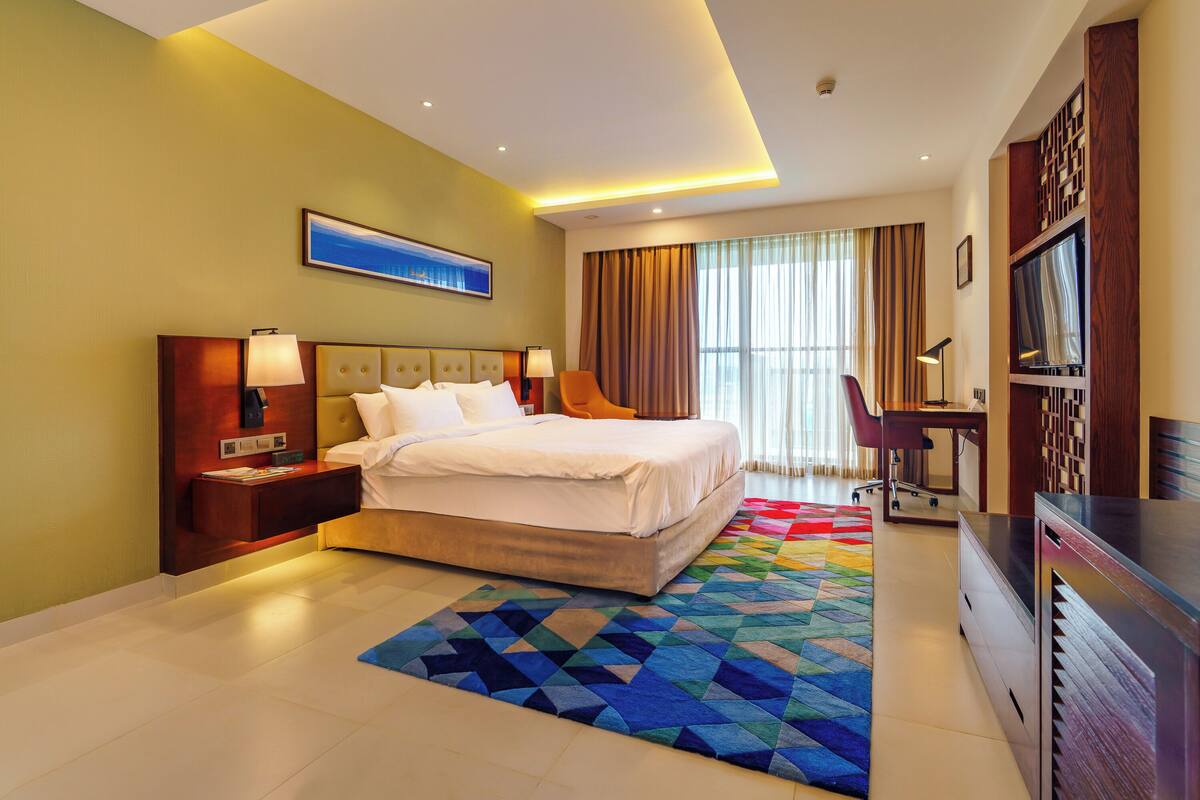 A hotel room with a bed, desk, chair, TV, and a large window with curtains.