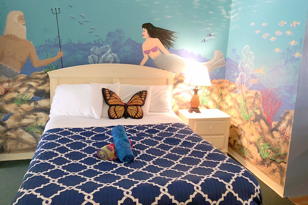 A bedroom with a mural of an underwater scene, a bed with a patterned bedspread, a pillow with a butterfly design, and a nightstand with a lamp.