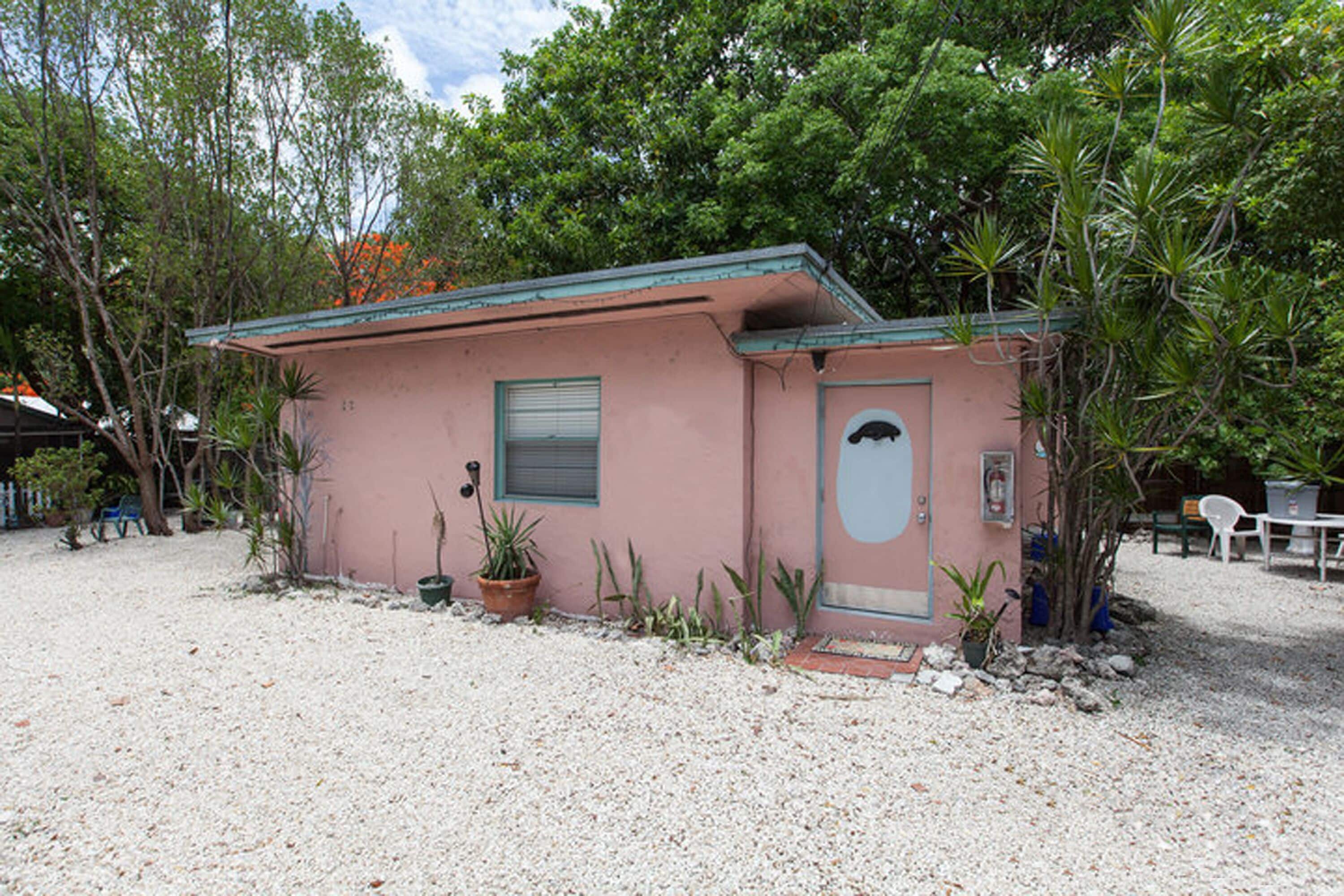 Un petit bâtiment rose avec un toit vert, entouré de gravier et de plantes en pot.