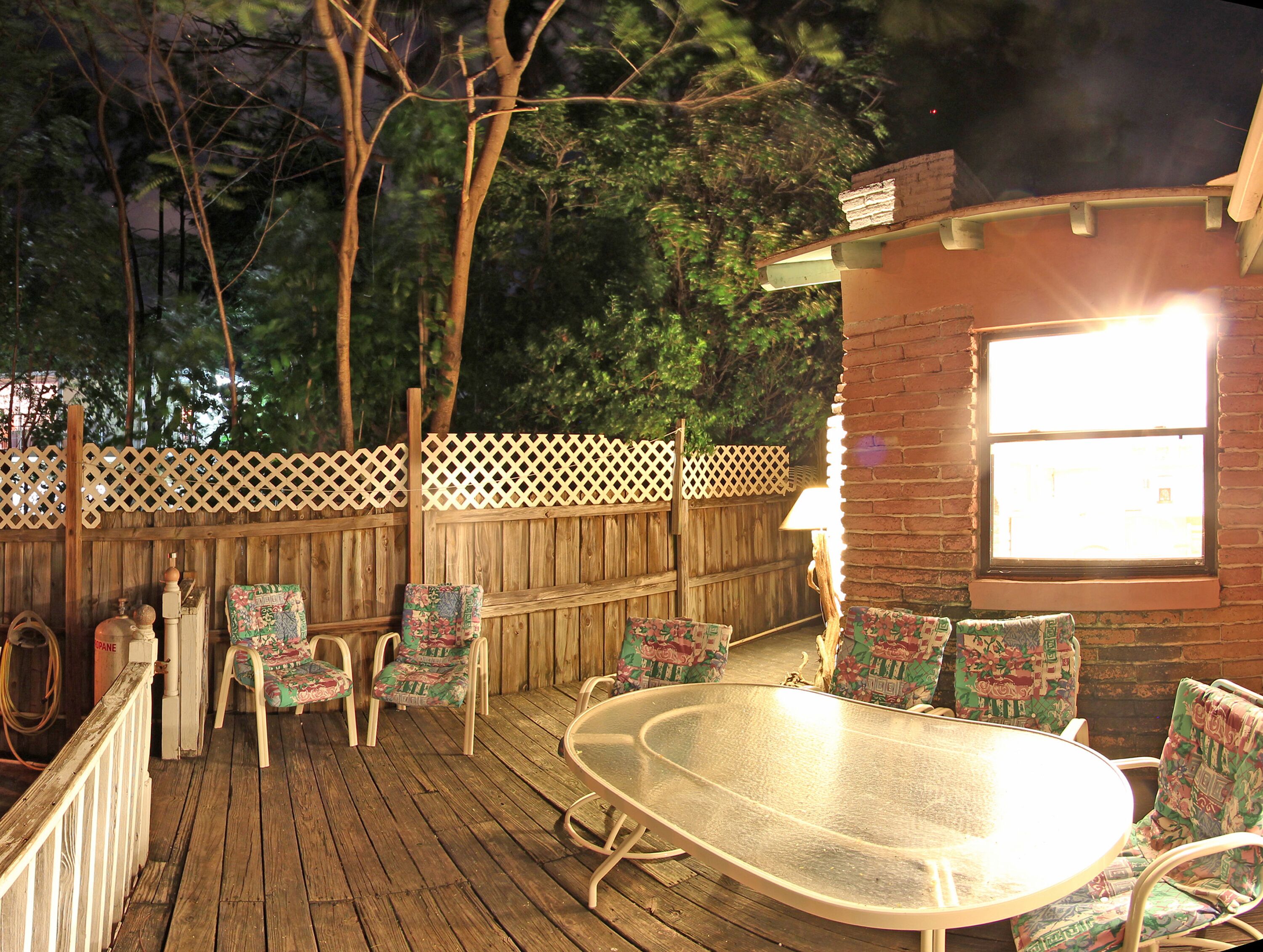 A wooden deck with a round table and chairs, a wooden fence, and a window with a view of trees.