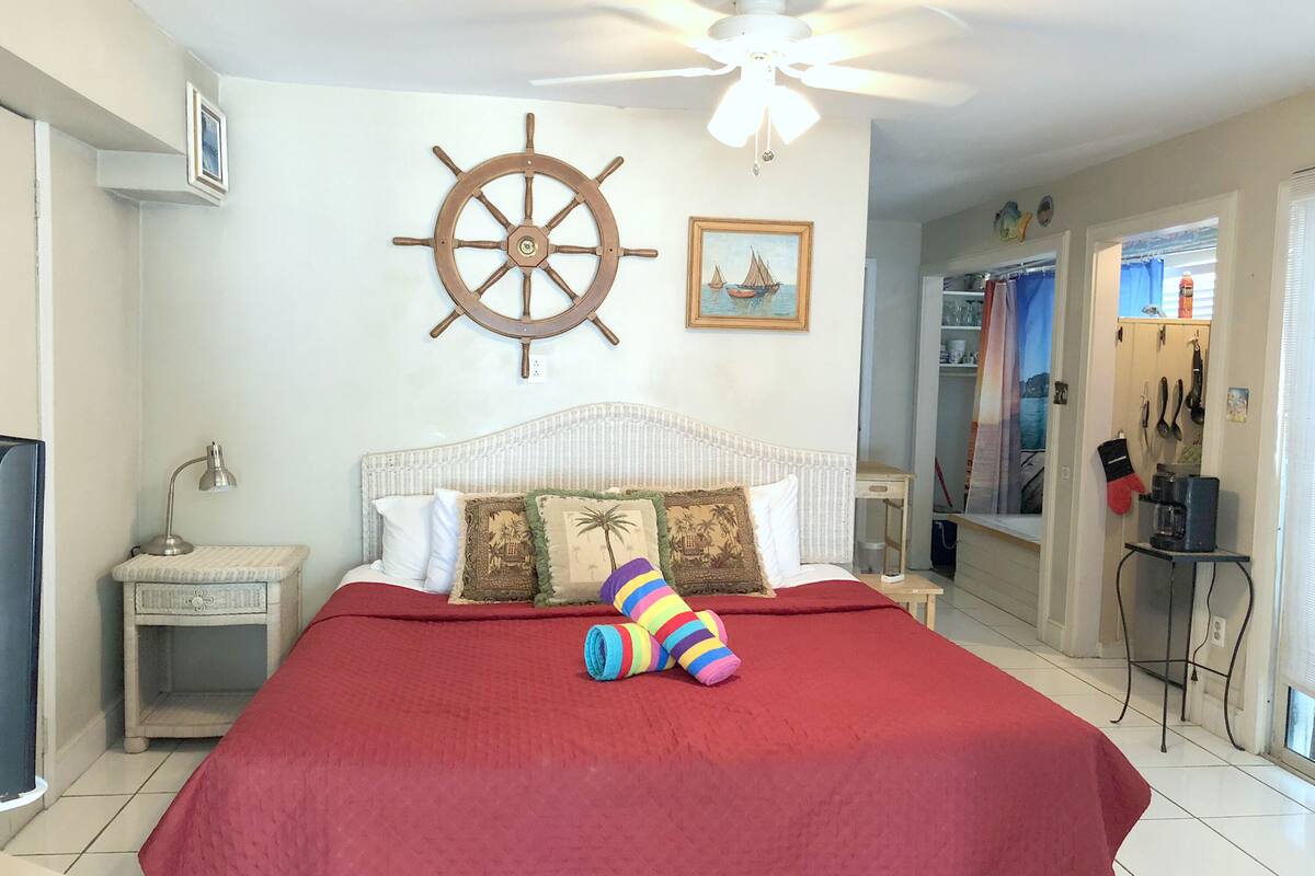 A bedroom with a bed, a ceiling fan, a ship's wheel decoration, and a painting of a boat.