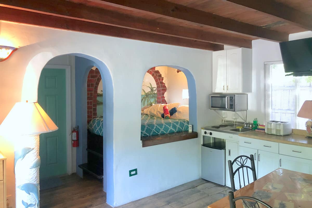 A compact kitchen with a microwave, toaster, and sink. There is a small dining table with chairs. The room has a wooden ceiling and a door leading to another room.